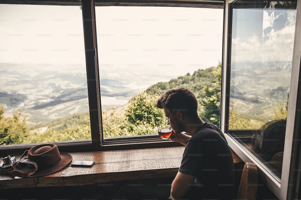세련된 힙스터 여행자는 숲과 산의 아름다운 풍경을 배경으로 유리 컵에 뜨거운 차를 마시고 있다. 산의 나무 창문에서 바라보는 전망. 텍스트를 위한 공간입니다. 여행하다