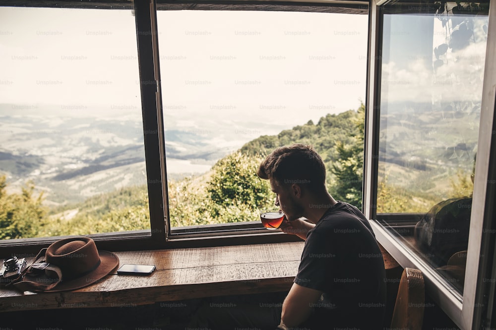 Stylish hipster traveler drinking hot tea in glass cup on background of  beautiful landscape of woods and mountains. View from wooden window on mountain. Space for text. Travel