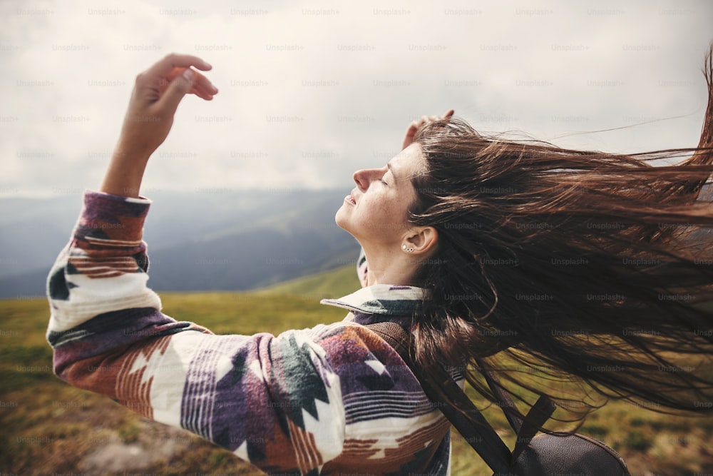Chica hipster con estilo con mochila y cabello ventoso en la cima de las montañas. Retrato de una joven feliz relajándose. Estado de ánimo despreocupado. Increíble momento atmosférico