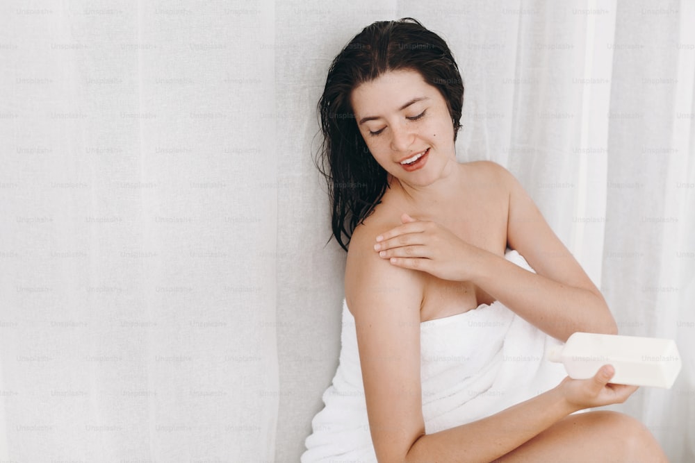 Young happy woman in white towel applying moisturizing cream on shoulder in bathroom. Skin and body care. Hand holding plastic bottle with lotion. Sexy woman relaxing, spa and wellness