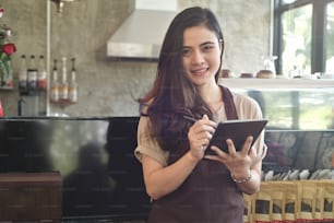 アジアの女性バリスタが笑顔でタブレットを使用してコーヒーショップのカウンターで注文を取る