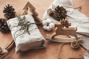 松ぼっくり、木、トナカイ、麻ひも、綿と木製のテーブルの上に緑の枝とリネン生地で包まれたスタイリッシュなクリスマス素朴な贈り物。シンプルエコはプラスチックフリーです。ゼロウェイストホリデー