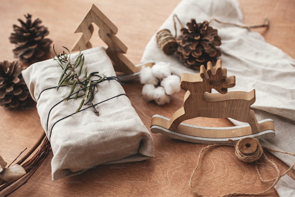 Stylish christmas rustic gift wrapped in linen fabric with green branch on wooden table with pine cones, tree,reindeer, twine, cotton. Simple eco presents plastic free. Zero waste  holidays