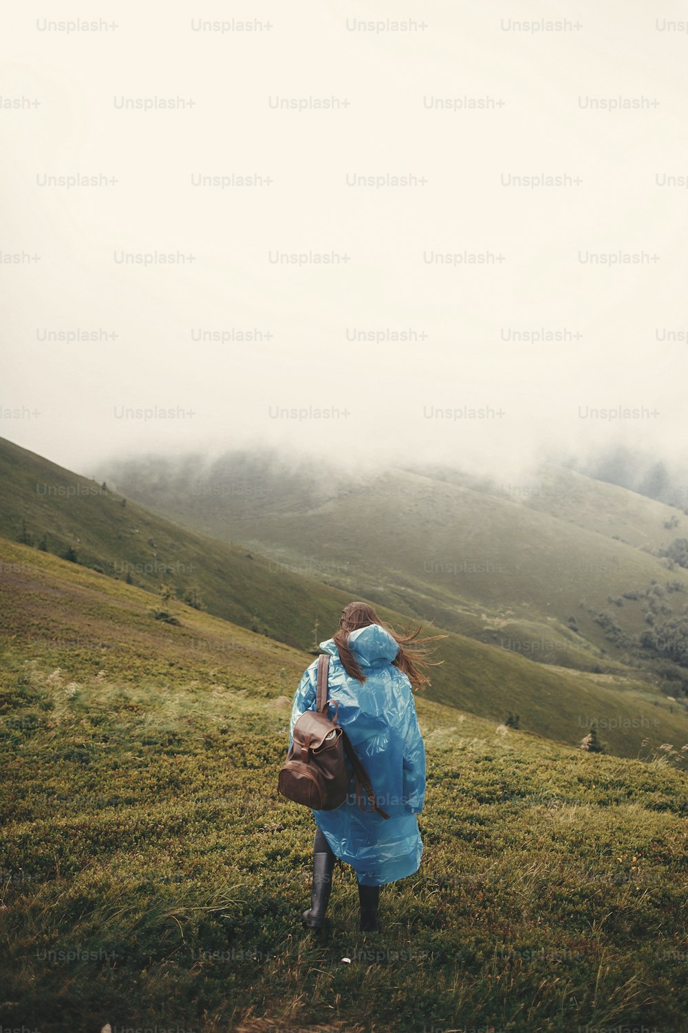 Garota hipster elegante em capa de chuva azul e com mochila olhando para colinas enevoadas no topo das montanhas. Mulher jovem viajante explorando montanhas. Conceito de viagem e desejo de viajar. Momento atmosférico