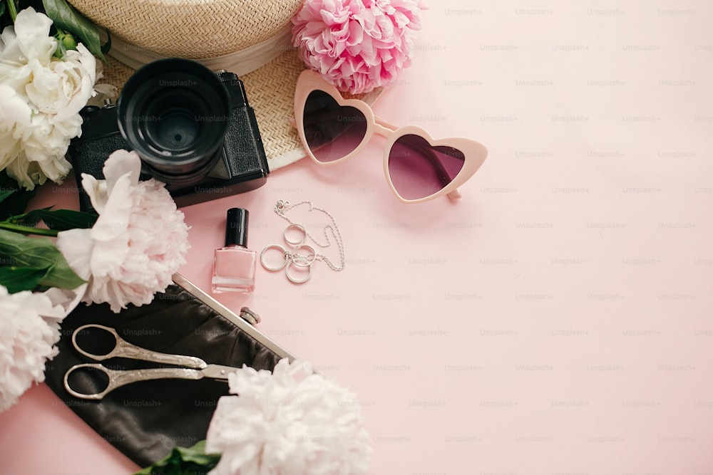 Stylish girly flat lay with pink peonies, photo camera, retro sunglasses, jewelry, nail polish, hat, purse on pastel pink paper with copy space. International Women's Day. Hello spring
