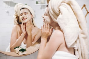 Young happy woman in towel making facial massage with  organic face scrub and looking at mirror in stylish bathroom. Girl applying scrub cream, peeling and cleaning skin. Skin Care
