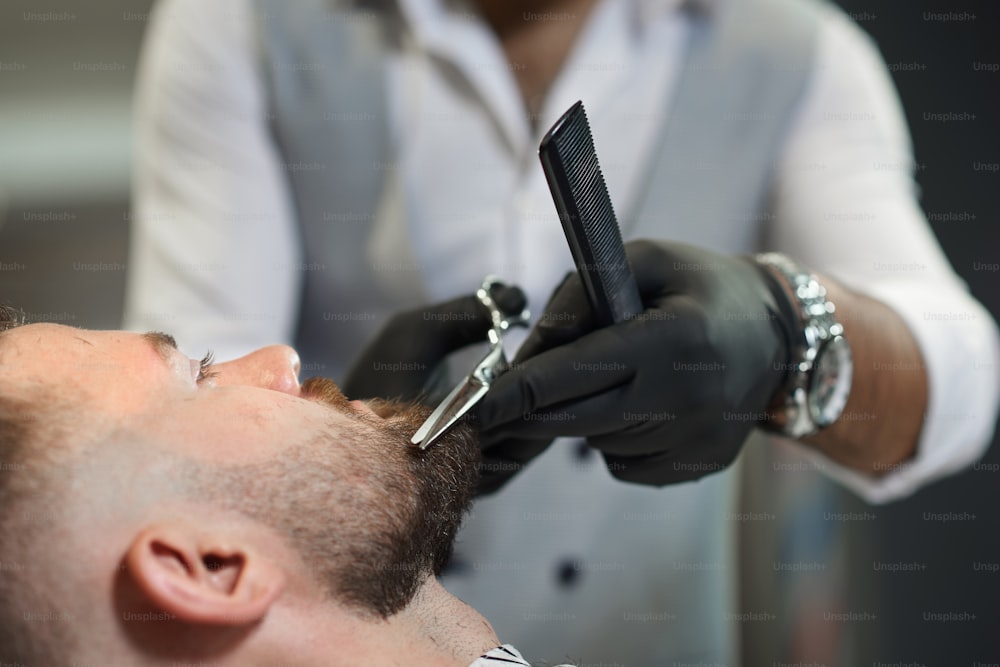 Primo piano del processo di taglio della barba del cliente maschio nel negozio di barbiere. Barbiere qualificato in guanti protettivi neri che tiene forbici e pettine in mano e corregge accuratamente la forma della barba.