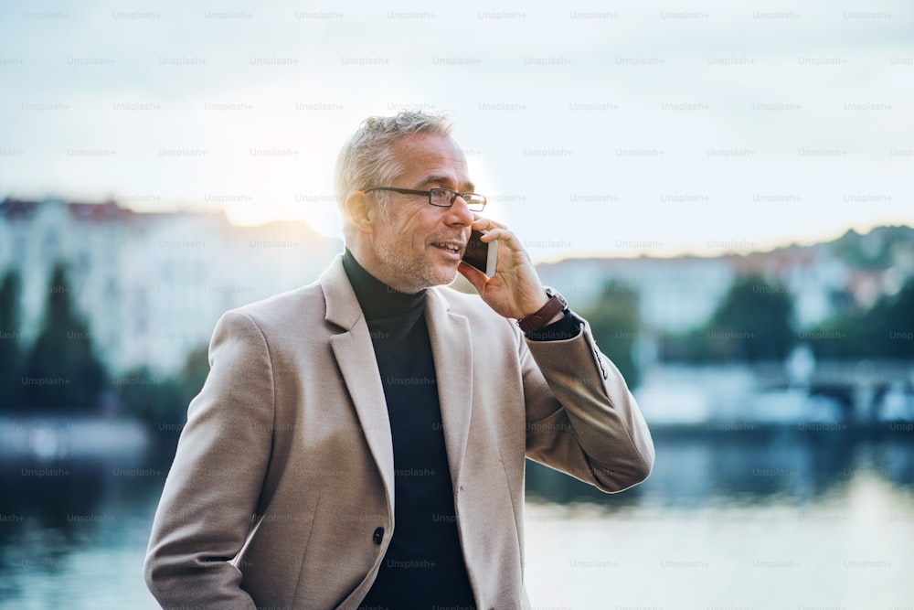 Mature businessman with smartphone standing by river in Prague city at sunset, making phone call.