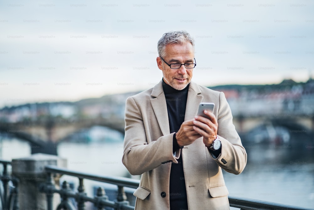 Mature handsome businessman with smartphone standing by river Vltava in Prague city at sunset, taking selfie. Copy space.