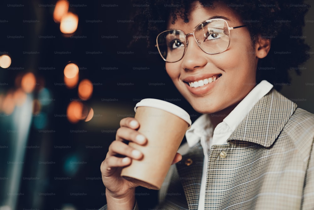 Portrait of cute woman in casual coat holding takeout beverage. She drinking coffee from brown cup of and smiling