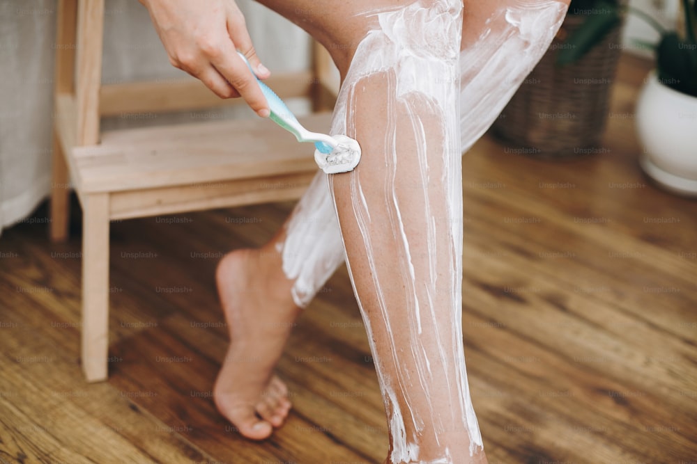 Hair Removal with depilation cream concept. Young woman in white towel applying shaving cream on her legs and holding plastic razor in home bathroom with green plants. Skin care