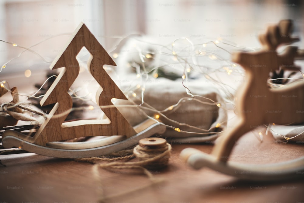 Rustikales Weihnachtsbaumspielzeug auf Holztisch auf dem Hintergrund von Lichtern, hölzernen Rentieren, Bindfaden, Geschenk in Leinen mit grünem Zweig, Zapfen. Frohe Weihnachten. Platz für Text. Einfache Öko-Geschenke