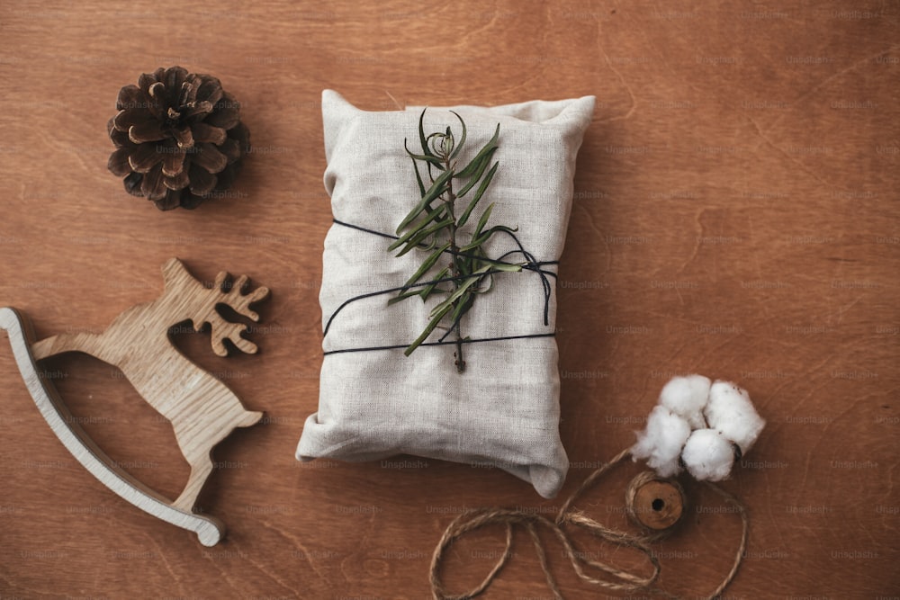 Stylish christmas rustic gift wrapped in linen fabric with green branch on wooden table with pine cone,reindeer, twine, cotton. Flat lay. Simple eco presents plastic free. Zero waste  holidays