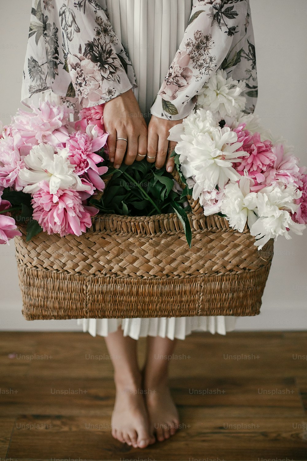 Boho girl holding pink and white peonies in rustic basket and standing barefoot on wooden floor. Stylish hipster woman in bohemian floral dress with peony flowers. International Womens Day