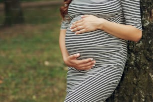 Beautiful pregnant woman in casual clothes, holding belly while posing in park closeup. maternity concept