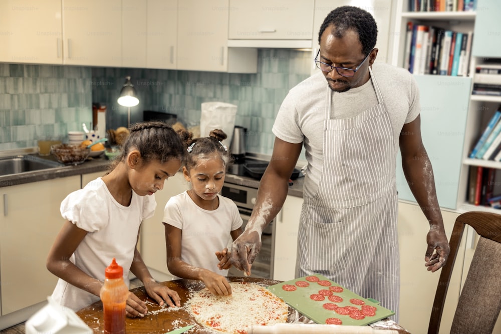 도움이되는 아버지. 줄무늬 앞치마를 입은 아버지는 어린 딸들이 살라미 소시지로 피자를 만드는 것을 돕고 있다