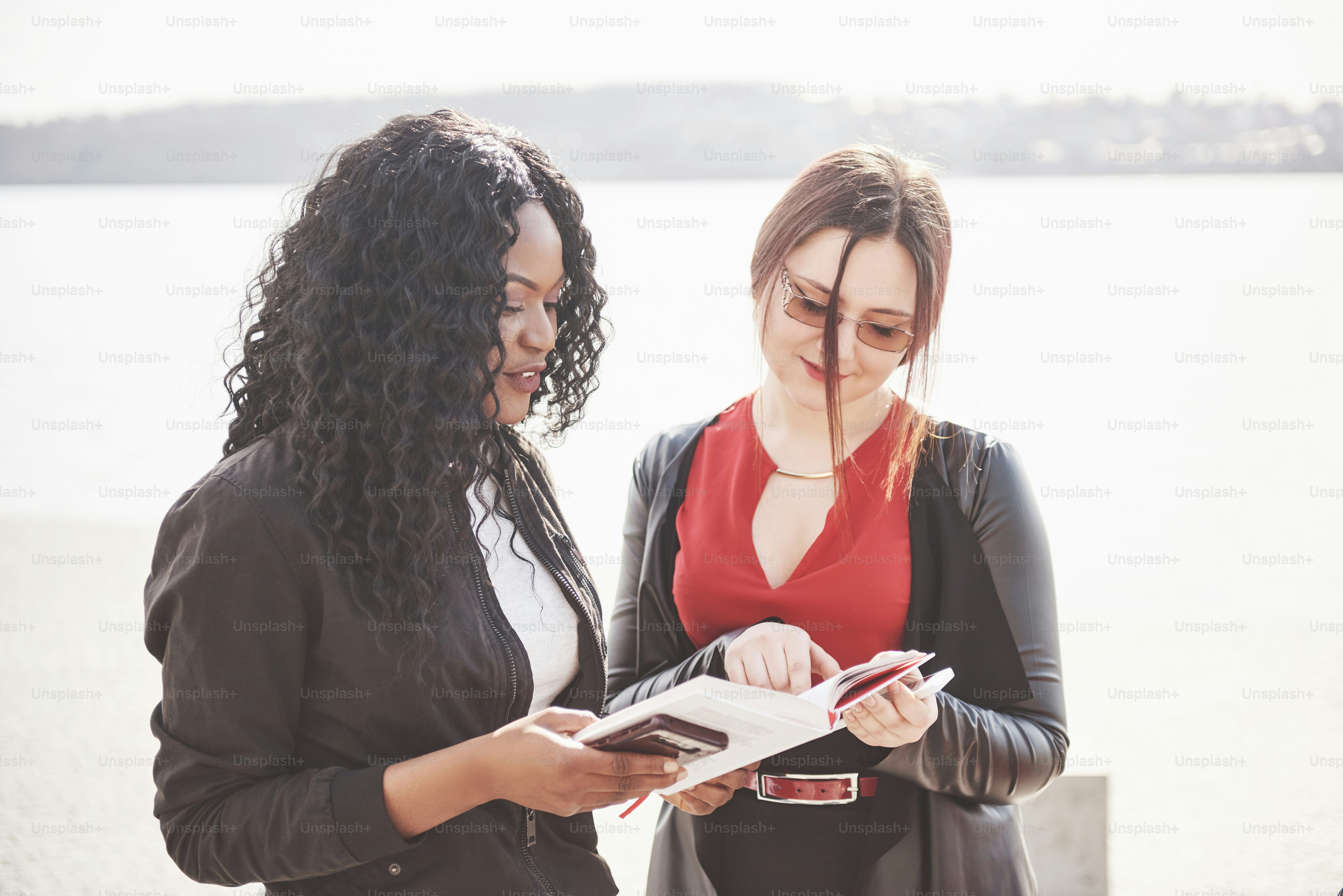 Two good friends of women are standing outside and reading a book.