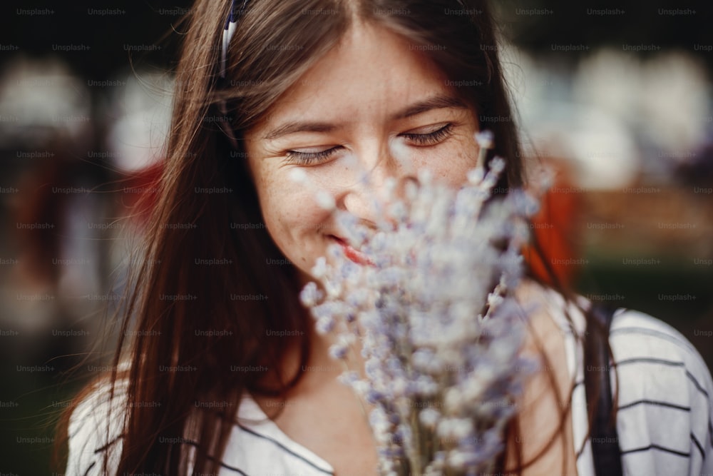 Beautiful stylish young woman holding amazing lavender flowers and smelling them in sunny outdoors. Happy hipster girl enjoying aroma of lavender bouquet. Calm atmospheric moment