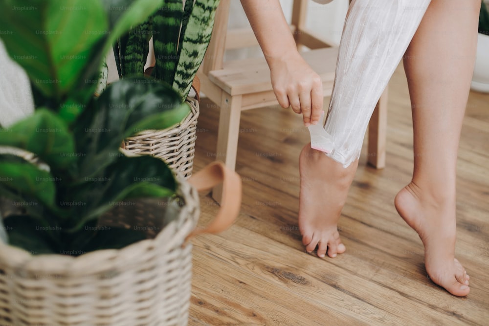 Young woman in white towel applying shaving cream on her legs in home bathroom with green plants. Skin care and wellness concept. Hand holding plastic razor on skin with cream