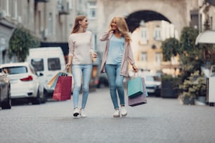 Full length portrait of charming young lady chatting with mother outdoors. They carrying colorful shopping bags while looking at each other and smiling