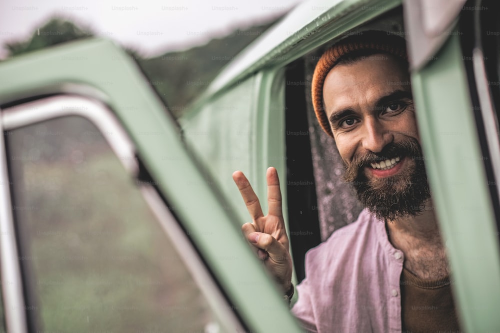 Concept of hipster lifestyle. Close up portrait of happy smiling caucasian male making peace gesture while sitting in retro van