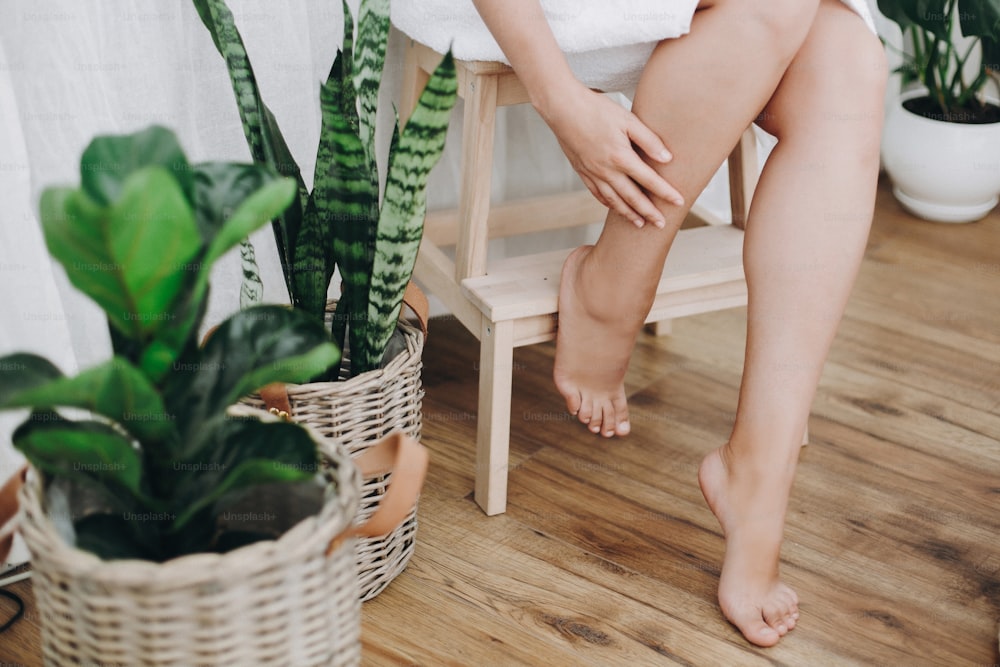 Skin care and wellness concept. Young woman in white towel sitting in bathroom with green plants and massaging legs. Legs soft skin after shaving close up.