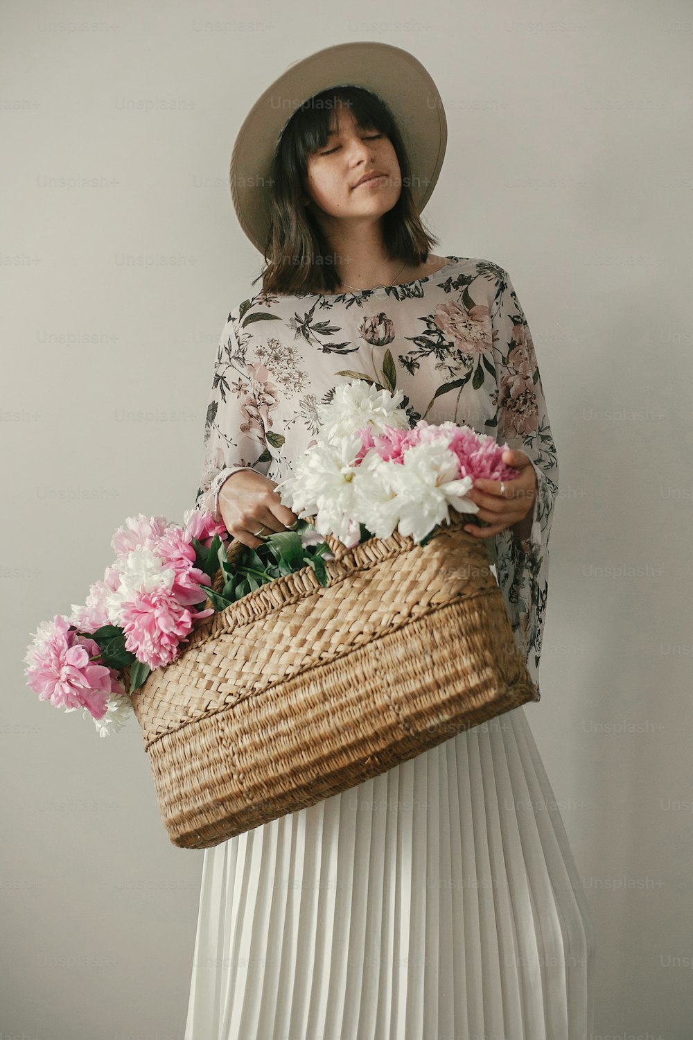 Boho girl in hat holding pink and white peonies in rustic basket. Stylish hipster woman in bohemian floral dress posing with flowers. International Womens Day. Countryside living