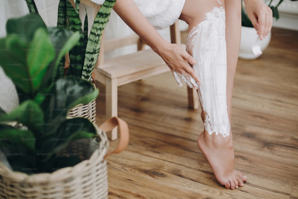 Young woman in white towel applying shaving cream on her legs in home bathroom with green plants. Skin care and wellness concept. Hand smearing moisturizer cream on skin