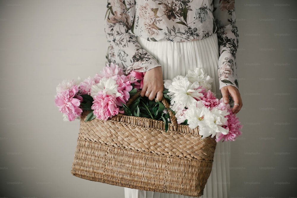 Boho girl holding pink and white peonies in rustic basket. Stylish hipster woman in bohemian floral dress gathering peony flowers. Happy mothers day. International Womens Day.