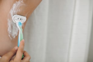 Hair Removal concept. Hand shaving armpit with depilation cream and plastic razor close up. Young woman epilating armpits closeup in home bathroom. Skin care. Copy space.