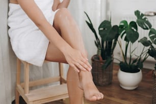 緑の植物のあるバスルームで髭を剃った後、足にクリームを塗る若い女性。スキンケアとウェルネスのコンセプト。柔らかい肌の結果のための保湿剤クリーム塗抹の足を持つ女の子の手