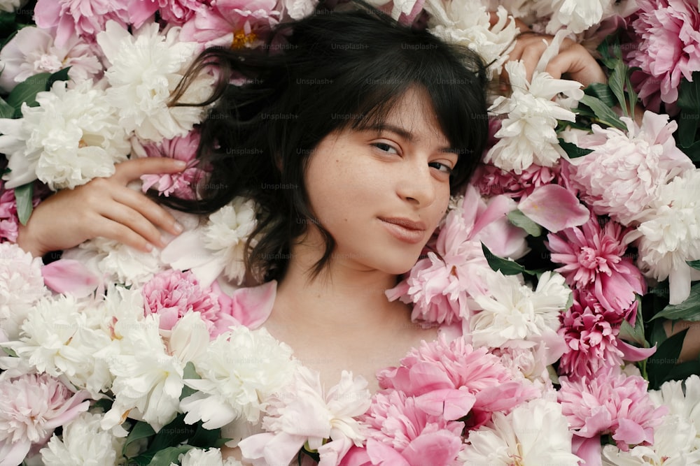 Porträt einer Boho-Frau mit natürlichem Make-up, die in Pfingstrosen liegt. Kreatives Blumenfoto. Aroma- und Spa-Konzept. Internationaler Frauentag. Schönes brünettes Mädchen in vielen rosa und weißen Blumen