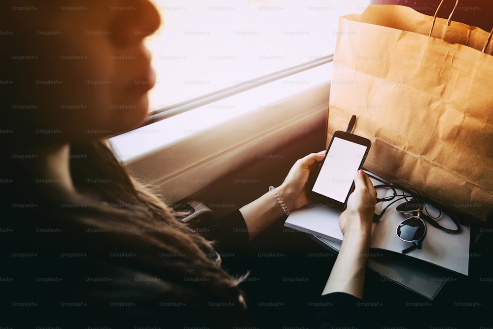 beautiful hipster girl traveling by train and holding phone. stylish happy woman looking at smartphone screen and smiling at window light in train. space for text