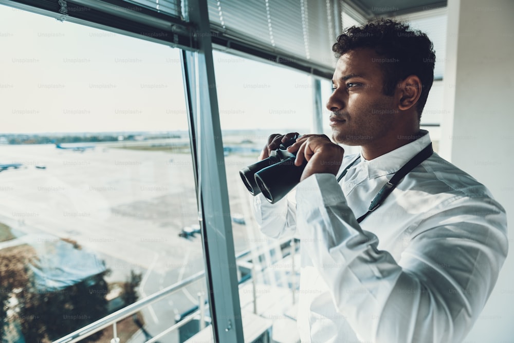 Center of dispatching maintenance. Waist up side on portrait of young specialist holding binocular glasses while staying near big window of navigation controller office
