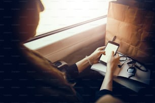 stylish hipster girl holding phone with headphones at window light in train. beautiful young woman looking at smartphone empty screen, bag and sunglasses on table. space for text