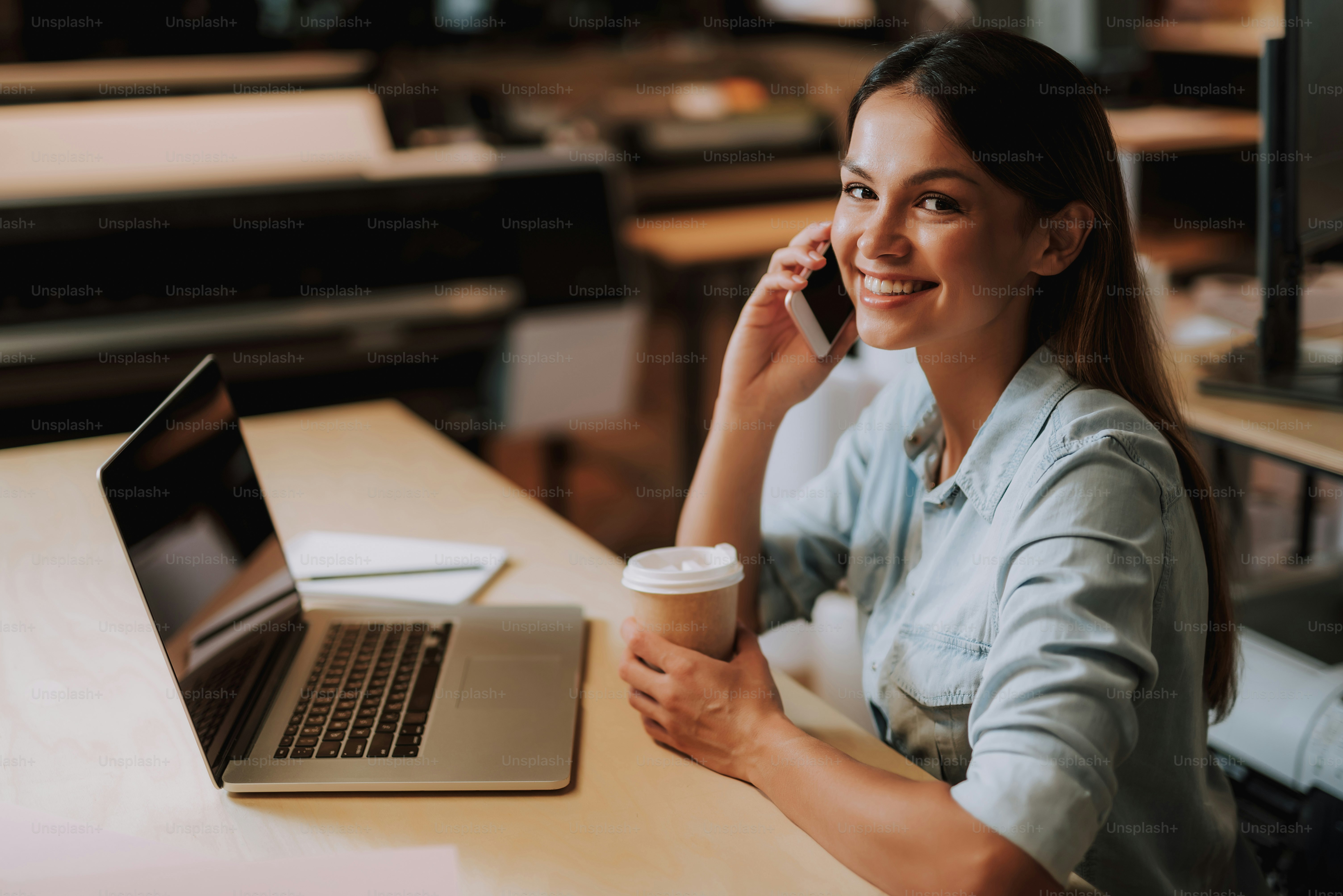 Portrait of beautiful girl with cup of coffee having phone conversation while sitting at office desk. She is looking at camera and smiling