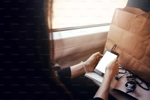 Stilvolles Hipster-Mädchen, das Telefon mit Kopfhörern bei Fensterlicht im Zug hält. Anreise mit dem Zugkonzept. Schöne junge Frau, die auf den leeren Bildschirm des Smartphones schaut. Platz für Text