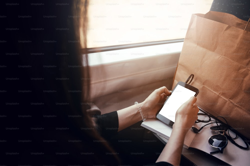stylish hipster girl holding phone with headphones at window light in train. travelling by train concept. beautiful young woman looking at smartphone empty screen. space for text