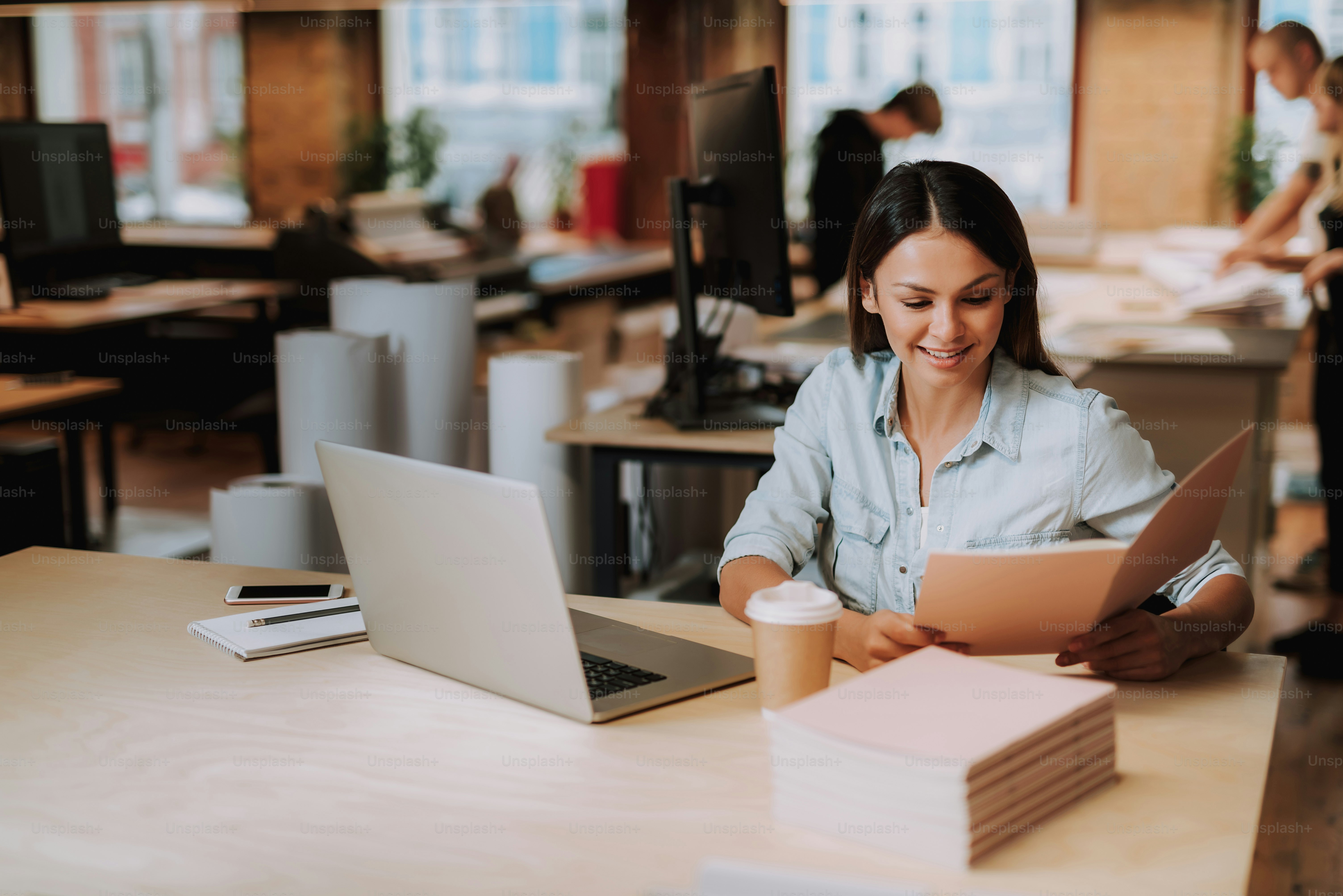 Portrait of beautiful woman reading documents while sitting at table with laptop, cellphone, cup of coffee and notebook