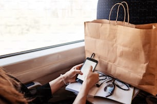 stylish hipster girl holding phone with headphones at window light in train. travelling by train concept. beautiful young woman looking at smartphone empty screen. space for text