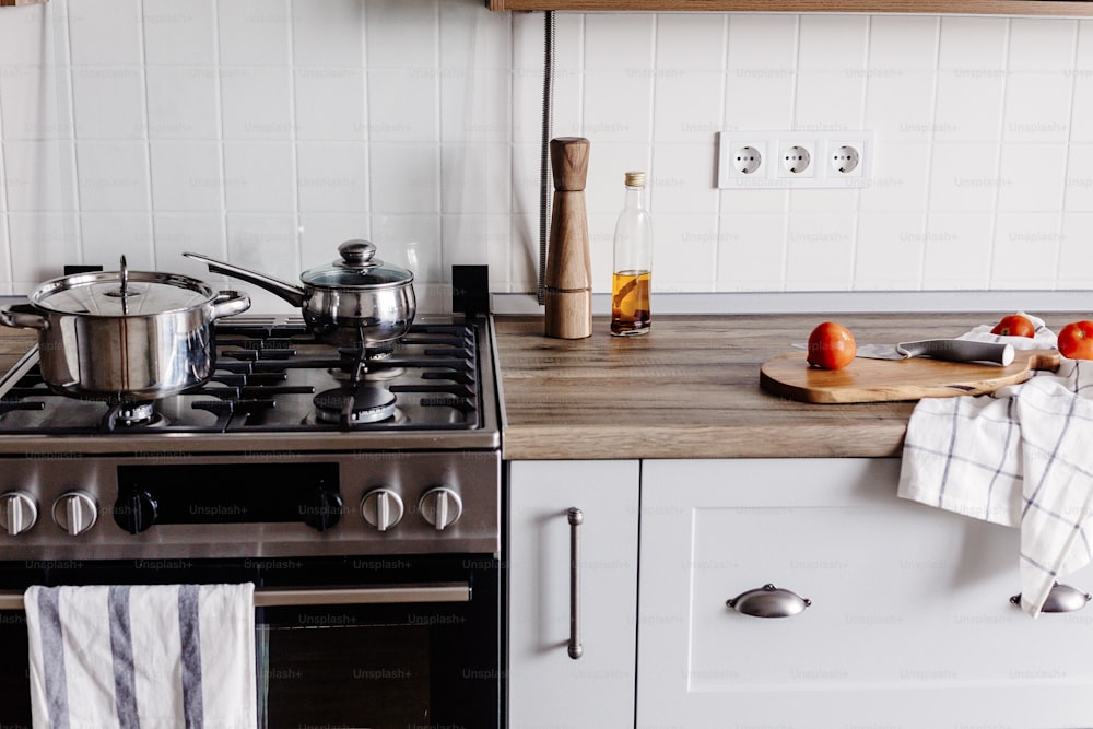 Cocinar alimentos en una cocina moderna con horno de acero, ollas, cuchillo en una tabla de cortar de madera con verduras, pimienta, especias, aceite sobre una mesa de madera. Comida casera. Muebles de cocina con estilo en color gris