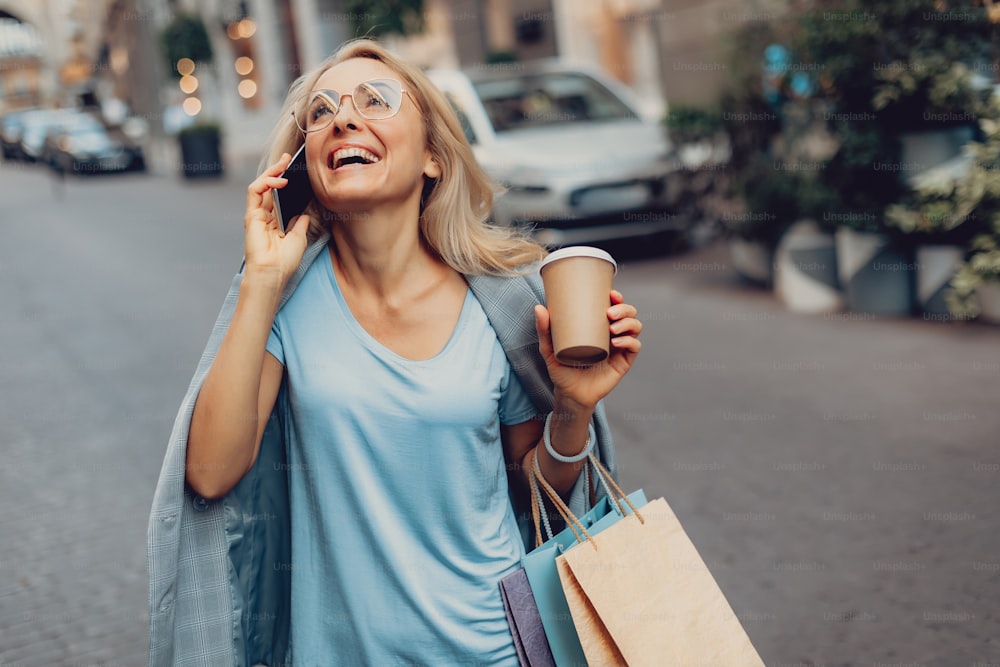 Waist up portrait of cheerful middle-aged lady in glasses talking on cellphone on the street. She is holding cup of coffee and shopping bags