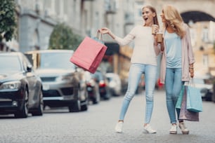 Retrato de corpo inteiro da menina bonita e sua mãe segurando sacolas de compras e xícaras de bebida quente. Mulheres olhando para o lado e sorrindo como se vissem algo excitante