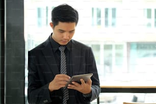 Confident young Asian businessman working on digital tablet while standing in office