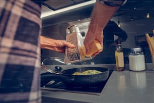 Fromage. Homme de grande taille portant une chemise à carreaux râpant du fromage sur l’omelette tout en cuisinant dans la cuisine
