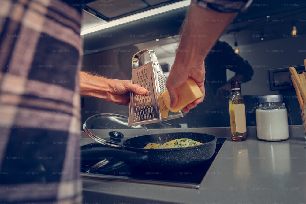 Cheese. Tall man wearing a checkered shirt grating cheese on the omelet while cooking in the kitchen
