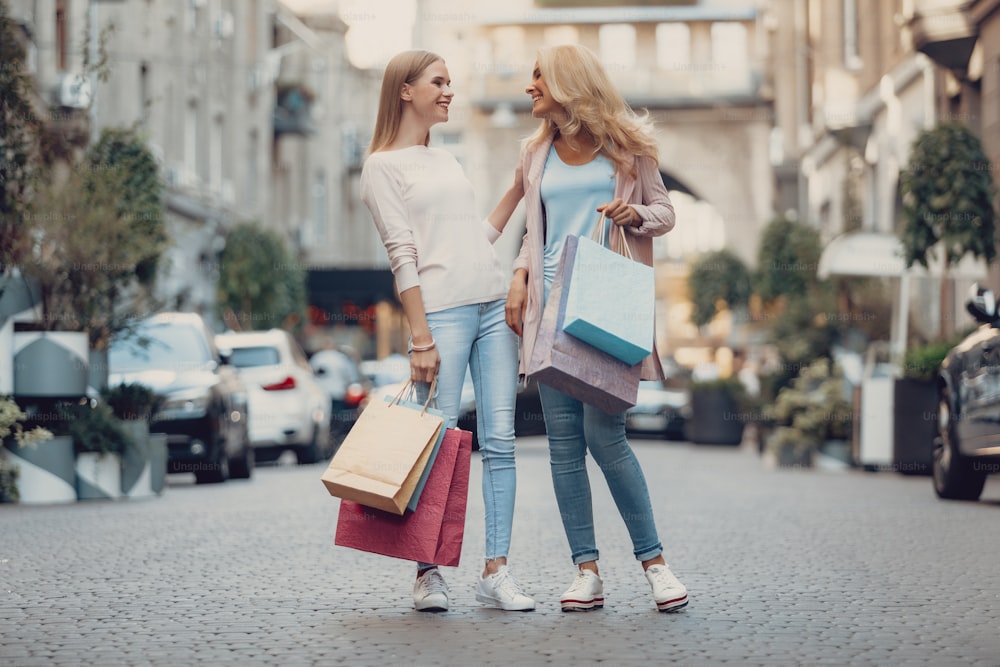 Full length portrait of beautiful girl touching shoulder of mother while looking at her with smile. Women holding colorful shopping bags