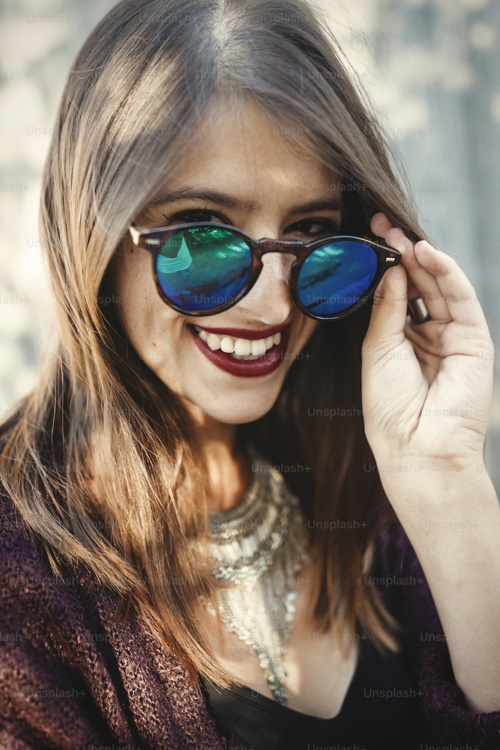 Stylish hipster girl smiling in sunny street on background of wooden wall. Portrait of boho girl in cool outfit and sunglasses posing in sunlight and shadow.  Summer vacation and travel
