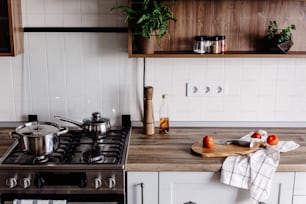 Cocinar alimentos en una cocina moderna con horno de acero, ollas, cuchillo en una tabla de cortar de madera con verduras, pimienta, especias, aceite sobre una mesa de madera. Comida casera. Muebles de cocina con estilo en color gris