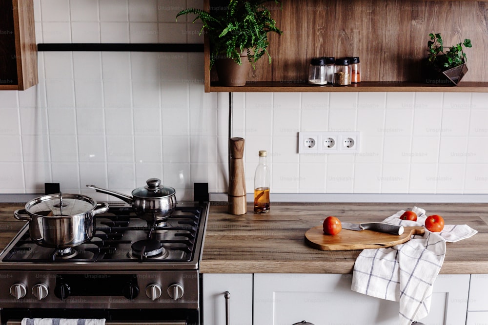Cocinar alimentos en una cocina moderna con horno de acero, ollas, cuchillo en una tabla de cortar de madera con verduras, pimienta, especias, aceite sobre una mesa de madera. Comida casera. Muebles de cocina con estilo en color gris